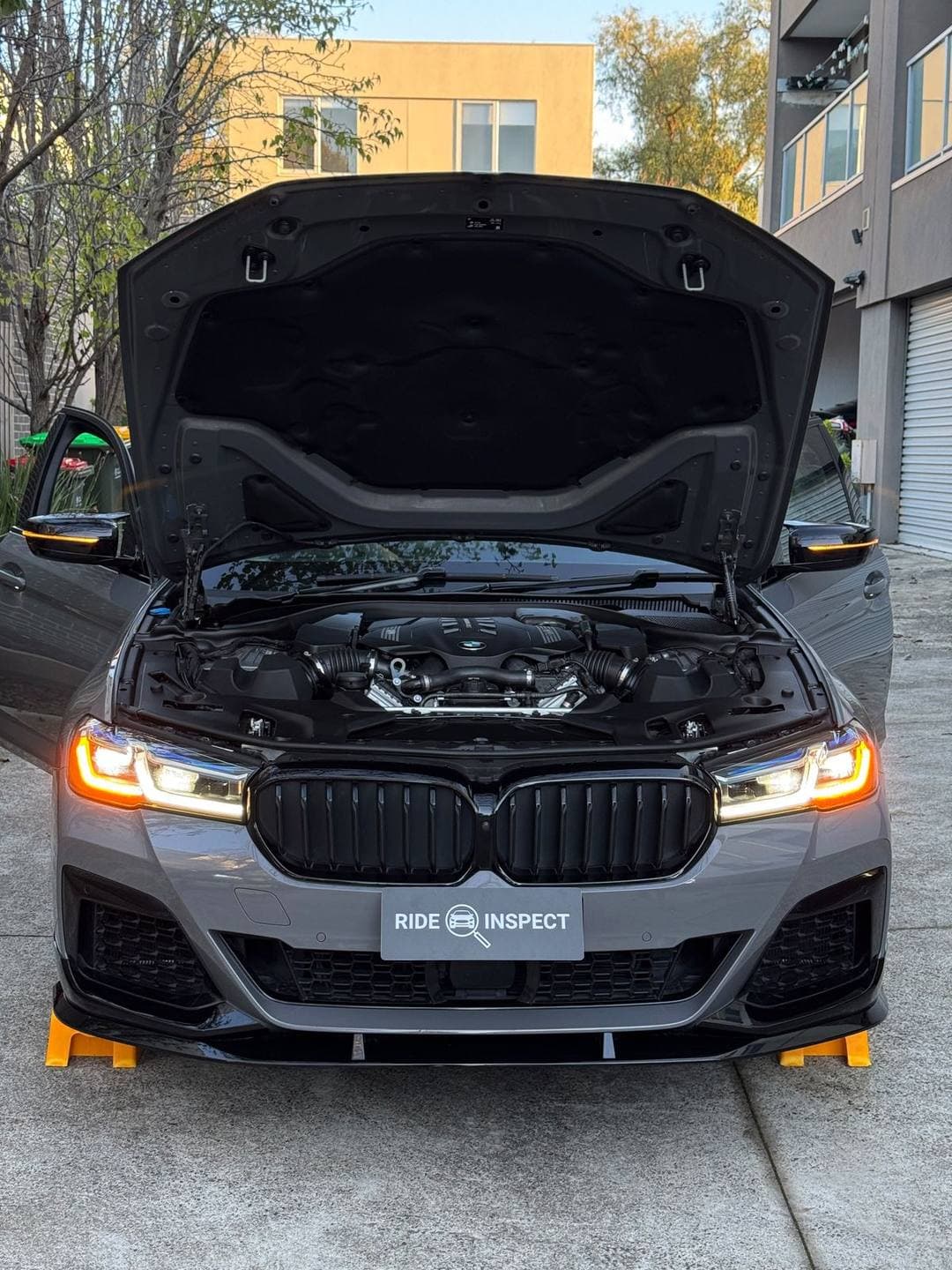 Grey BMW 5 Series from the front, bonnet up and headlights on, in an apartment driveway
