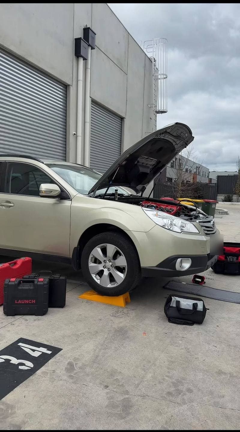 Gold Subaru Outback with the bonnet up in a car park, tool cases and bags around it