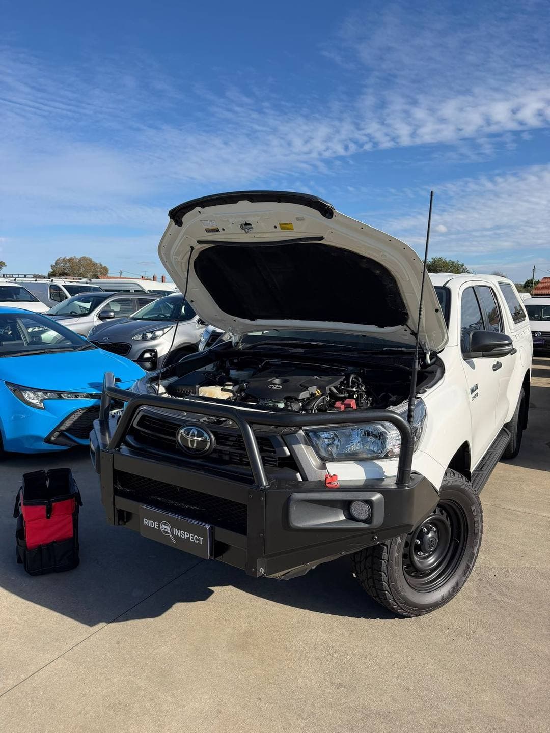 White Toyota HiLux ute with a bullbar and snorkel, bonnet up in a car yard, with the mechanic's tool bag beside it